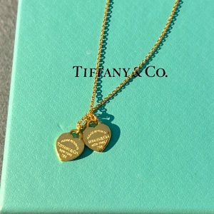 Guldigt halsband Tiffany hjärtan -  guld med två hjärtformade berlocker. Klassisk kedja och snygga detaljer med ingraverad text på berlockerna. Perfekt statement-piece för dig som gillar lyxiga smycken med ikonisk design. Utan låda