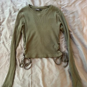 Olivgrön ribbad topp med snörning - Säljer en olivgrön långärmad topp från FB Sister i ribbad bomull. Tröjan har croppad passform och snygg snörningsdetalj i sidorna som ger en trendig look. Perfekt till jeans eller kjol för en chill vibe.