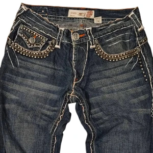 Laguna Beach jeans med nitar och brodyr - Unika mörkblå jeans från Laguna Beach med kontrastsömmar, silverfärgade nitar och broderade detaljer på fickorna. Snygga knappar med döskallemotiv och coola fickor med gul brodyr bak. Perfekta för dig som vill sticka ut med din stil.