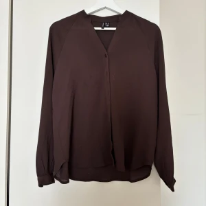 Brun blus med v-ringning från Vero Moda - Snygg brun blus från Vero Moda i storlek XS. Modellen har v-ringning, knappar framtill och långa ärmar med enkel manschett. Tunt och lätt material som ger ett luftigt intryck. Perfekt att styla med jeans eller kjol för en clean look.