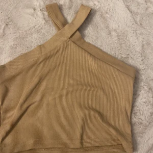 Beige ribbad linnetopp med korsade band - Snygg beige ribbad linnetopp med korsade axelband framtill. Toppen är ärmlös och har en tight passform som framhäver siluetten. Perfekt för dig som gillar stilrena och trendiga plagg med det lilla extra i designen. Med en fläck.