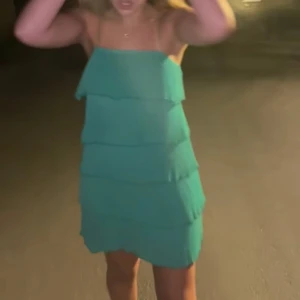 Grön volangklänning med smala axelband  - Säljer en grön kortklänning med flera volanglager och smala axelband. Klänningen har en luftig passform och är perfekt för varma dagar. Enkel men ändå iögonfallande design som ger en fräsch look.