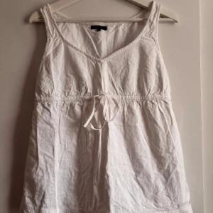 Vit Linnetopp Tommy Hilfiger - Supersöt vit linnetopp från Tommy Hilfiger med broderade blommor, v-ringning och knytband i midjan. Toppen är ärmlös och har dragkedja i sidan för enkel påklädning. Perfekt för varma dagar och ger en fräsch sommarkänsla. Skulle säga att storleken motsvarar S/M men lite mer S! Använd ett par gånger men inga defekter🤗