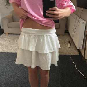 Vit volangkjol med resårmidja - Superfin vit kortkjol med dubbla volanger och bred resår i midjan. Kjolen är lätt och luftig, perfekt för varma dagar. Materialet känns som bomull eller linne och ger en skön vibe. Enkel att matcha med både t-shirt och crop top.