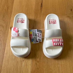 Hello Kitty vita sandaler med platåsula - Supergulliga vita sandaler med platåsula och öppen tå, dekorerade med Hello Kitty-logga och figur i rosa och vitt. Perfekta för dig som älskar kawaii-stil och vill sticka ut med något unikt. Tillverkade i syntetmaterial och riktigt bekväma att glida runt i. De passar storlek 36-37.