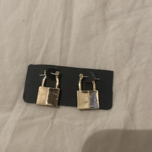 Guldiga örhängen med hänglåsdesign - Coola örhängen i form av små hänglås i guldigt metallmaterial. De har en unik och edgy känsla som verkligen sticker ut. Perfekta för dig som vill ha något annorlunda och trendigt i smyckeskrinet.