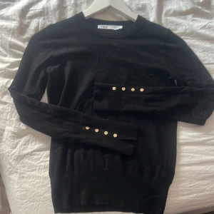 Svart tröja med guldknappar från Zara - Svart långärmad tröja från Zara med snygga guldknappar på ärmarna. Enkel och stilren design med rund halsringning. Storlek S