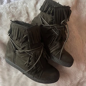 Olivgröna mockaboots med fransar - Snygga gröna khaki kilklack boots i mocka med coola fransdetaljer runt hela skaftet och snörning framtill. Skorna har en platt sula med klack och rund tå, perfekt för dig som vill sticka ut med en boho-vibe. Passar till både jeans och klänning för en trendig look.