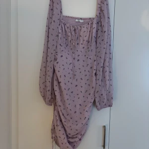 Ljuslila blommig klänning från NA-KD - Supersöt ljuslila kortklänning från NA-KD med små blommor i lila och grönt. Klänningen har långa puffärmar, smock upptill, knytning vid halsen och snygg rynkdetalj längs sidan. Perfekt för dig som gillar romantisk och trendig stil.