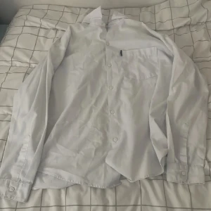 Vit skjorta från Free Wear 158/164 - Klassisk vit skjorta från Free Wear i storlek 158/164. Skjortan har lång ärm, bröstficka och knäppning framtill. Perfekt till skjorta-looken med krage och enkel stil. Materialet känns som bomull och passar till många olika outfits.