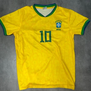 Brasilien Neymar Jr. 10 fotbollströja - Gul Brasilien fotbollströja med gröna detaljer, Neymar Jr. och nummer 10 på ryggen. Klassisk V-ringning med blå och grön kant, samt CBF-emblem på bröstet. Tillverkad i lätt och ventilerande material, perfekt för match eller träning.