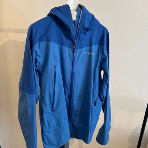 Blå vindjacka från Norrøna med huva - Snygg blå vindjacka från Norrøna med justerbar huva och flera dragkedjeförsedda fickor. Jackan har en sportig look, hel dragkedja framtill och är perfekt för utomhusaktiviteter. Materialet är lätt och känns smidigt, perfekt för dig som gillar äventyr.