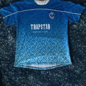 Blå Trapstar fotbollströja - Snygg blå fotbollströja från Trapstar med vit logga och texten 'IT'S A SECRET' på bröstet. Tröjan har ett mönster över hela ytan, vita detaljer och nummer 22 på ryggen. Tillverkad i ett lätt och ventilerande material, perfekt för match eller träning.