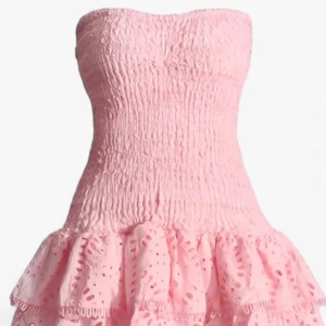 Rosa axelbandslös kortklänning - Supersöt rosa kortklänning med smockad överdel och volangig kjol. Axelbandslös modell med broderade detaljer och hålmönster i kjolen. Perfekt för sommarens alla tillfällen när du vill känna dig extra fin.