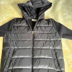 Svart Moncler cardigan med huva - Svart cardigan från Moncler med stickade ärmar och huva. Jackan har dragkedja framtill, två fickor med knappstängning och Moncler-logga på ärmen. Ärmarna har röd, vit och svart rand längs axeln i en stilren bladning. Oanvänd. Säg till för fler bilder eller funderingar☺️
