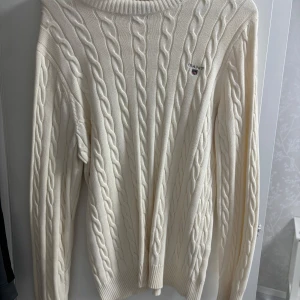 Vit kabelstickad tröja från Gant - Kabelstickad tröja från Gant i mjuk vit bomull. Klassisk design med rund hals, ribbade muddar och diskret Gant-logga på bröstet. Perfekt för dig som gillar en clean och tidlös look med lite preppy känsla. Dem är av storlek L på under sellout tröjan och gant är storlek sS