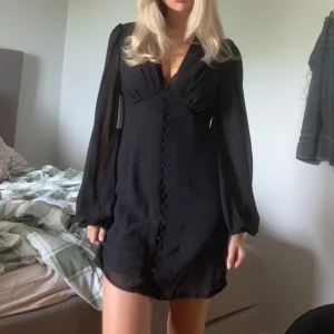 Svart kort klänning - Snygg svart kort klänning med v-ringning och långa ärmar, liknar en adoore klänning i modellen. Klänningen har dekorativa knappar framtill och ett lätt, flowy tyg som ger en elegant känsla. Perfekt för dig som gillar stilrena och trendiga plagg.