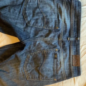 Blå jeans från Lee bootcut - Säljer ett par klassiska blå jeans från Lee med bootcut-modell, knappt använda och en låg midja. Jeansen har Lee-logga på bakfickan. Tillverkade i jeansmaterial med en mörkblå tvätt och kontrastsömmar. Mått: midja - 40cm, innerbenet- 74,5cm.