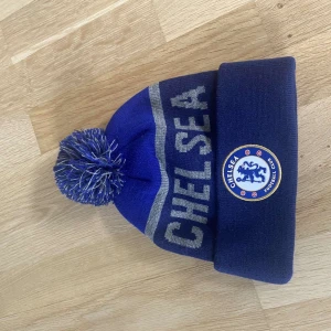 Chelsea FC blå mössa med tofs - Blå stickad mössa med Chelsea FC-logga framtill och stor text 'CHELSEA' i grått. Mössan har en bred uppvikt kant och en blå-vit tofs på toppen. Perfekt för dig som vill visa ditt stöd för laget under kalla dagar.