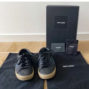 Svarta Saint Laurent sneakers i skinn - Svarta sneakers från Saint Laurent i skinn med handskriven logga i blått på sidan. Klassisk låg modell med snörning och beige gummisula. Snygg och stilren design som passar till det mesta. Levereras med originalkartong och dustbags.