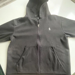 Polo Ralph Lauren zip hoodie - Svart zip hoodie från Polo Ralph Lauren med dragkedja framtill och klassisk broderad logga på bröstet. Tröjan har huva och är tillverkad i mjuk bomullsmix. Perfekt för en chill och stilren look.