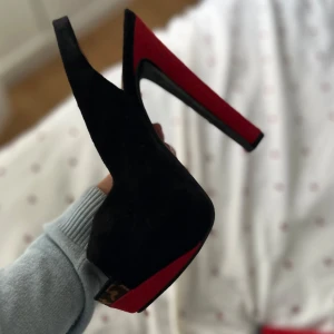 Svarta och röda pumps med leoparddetalj - Snygga pumps i svart mocka med röda detaljer på klacken och sulan. Skorna har öppen tå, hög klack och leopardmönstrad insida vid hälen. Perfekta för dig som vill sticka ut med en cool och elegant look.