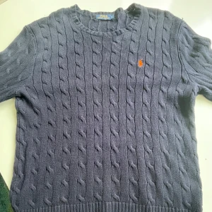 Polo Ralph Lauren kabelstickad tröja - Kabelstickad mörkblå tröja från Polo Ralph Lauren med klassisk orange broderad logga på bröstet. Tröjan har rund halsringning och långa ärmar. 