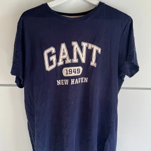 Marinblå GANT t-shirt med tryck - Snygg marinblå t-shirt från GANT med klassiskt tryck 'GANT 1949 New Haven' på bröstet. T-shirten har rund hals, korta ärmar och är gjord i mjuk bomull. Perfekt för en avslappnad och sportig look.