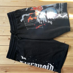 Svarta shorts Black Squad XL fjärilstryck - Svarta shorts från Black Squad i storlek XL med coolt tryck av en blå fjäril och eld, samt vit text längs benen. Mjuk resår i midjan med snörning och bakficka. Perfekta för chill dagar eller streetwear vibes.