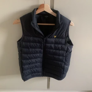 Mörkblå dunväst från Lyle & Scott - Snygg mörkblå dunväst från Lyle & Scott med gul logga på bröstet. Västen har quiltad design, hög krage och dragkedja framtill. Perfekt att slänga över en hoodie för extra värme. Klassisk och stilren look. Stl 12-14 år. 
