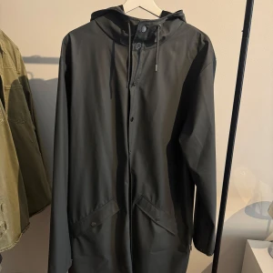 Svart regnjacka från Rains, storlek L - Snygg svart regnjacka från Rains i storlek L. Jackan har huva med snörning, tryckknappar framtill och två stora fickor med lock. Perfekt för regniga dagar och har en clean, minimalistisk look som funkar till allt.