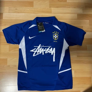 Blå Brasil Nike x Stüssy fotbollströja M - Snygg blå Brasil fotbollströja från Nike i samarbete med Stüssy. Tröjan har korta ärmar, vit Stüssy-logga på bröstet, CBF-emblem och vita detaljer på sidorna. Tillverkad i Dri-FIT material som andas, perfekt för fotboll eller streetwear. Ny med tags!