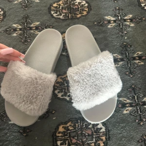 Grå fluffiga sandaler med päls - Supermysiga grå sandaler med bred rem i mjuk fuskpäls. Platt sula och öppen tå gör dem perfekta för chill dagar hemma eller när du vill ha något extra comfy. Snygg minimalistisk look med en touch av lyx tack vare det fluffiga materialet.