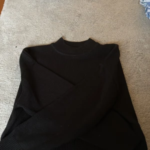 Svart stickad polotröja i storlek S - En svart stickad polotröja i storlek S med ribbade detaljer vid hals, ärmslut och nederkant. Tröjan har lång ärm och en klassisk, enkel design som är lätt att matcha med olika outfits.