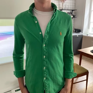 Grön skjorta från Ralph Lauren  - Snygg grön skjorta från Ralph Lauren i storlek S. Klassisk button-down med orange broderad logga på bröstet. Tillverkad i lätt bomullsmaterial, perfekt för dig som gillar stilrena och färgstarka plagg. Långärmad och med knappar hela vägen.