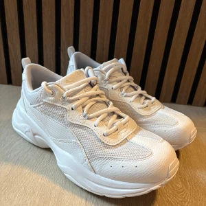 Vita sneakers från Puma - Snygga vita sneakers från Puma i storlek 40. Skorna har chunky sula, mesh och skinnpaneler samt klassisk snörning. Endast använda en weekend. Passade inte mina fötter. Har haft andra innersulor i så de är helt oanvända.