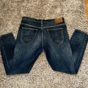 Mörkblå Lee Riders jeans - Snygga mörkblå Lee Riders jeans med klassisk femficksdesign och slitna detaljer framtill. Jeansen har kontrastsömmar, läderpatch med Lee-logga bak och coolt tryckt innerfoder. Perfekta för dig som gillar en tidlös och avslappnad look.