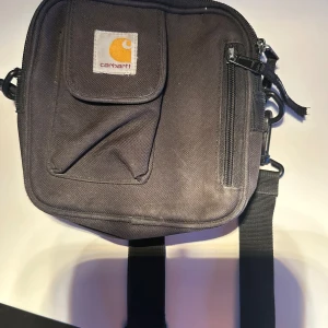 Svart axelväska från Carhartt - Säljer en svart axelväska från Carhartt med justerbar rem, praktisk framficka med lock och dragkedja samt extra fack framtill. Väskan är fyrkantig och tillverkad i slitstark polyester, perfekt för att bära dina viktigaste grejer.
