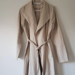 Beige trenchcoat från ONLY, stl S - Snygg beige trenchcoat från ONLY med klassisk krage och bälte i midjan. Jackan har långa ärmar och en stilren, rak passform. Perfekt för dig som vill ha en clean och tidlös look. Materialet känns mjukt och lätt, perfekt för vår och höst.  Helt & fint skick dock tvättludd 