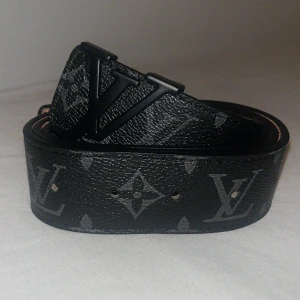 Svart Louis Vuitton skärp i skinn - Snyggt svart Louis Vuitton bälte i skinn med klassiskt monogrammönster och stor LV-spänne i svart metall. Bältet har en lyxig känsla och är perfekt för att lyfta vilken outfit som helst. Insidan är ljusbrun och bältet är tillverkat i Frankrike.