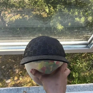 Svart keps från Gucci med GG-mönster - Snygg svart keps från Gucci med klassiskt GG-mönster över hela kepsen. Kepsen har böjd skärm och mesh på insidan för extra ventilation. Perfekt accessoar för att lyfta din outfit med en lyxig touch.