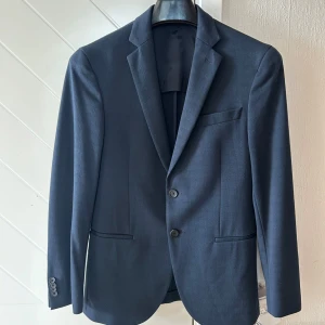 Mörkblå kavaj från Hugo Boss - Snygg mörkblå kavaj från Hugo Boss med klassisk passform. Kavajen har två knappar framtill, smala slag och tre knappar vid ärmslutet. Tillverkad i ett mjukt material som ger en stilren look. Perfekt för dig som vill ha en tidlös och elegant stil. Kavajen är fodrad i axelparti och framsidan, ofodrad i ryggpartiet. Mycket fint skick, inte mycket använd. Storlek 46