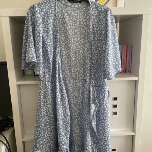 Blåvit omlottklänning med leopardmönster - Supersöt blåvit omlottklänning  med korta ärmar och trendigt leopardmönster. Klänningen har volangdetalj nertill och knytband i midjan för en snygg passform. Perfekt för dig som vill sticka ut med en cool och fräsch look. 