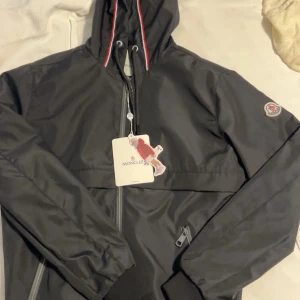 Svart vindjacka från Moncler - Svart vindjacka från Moncler med huva och dragkedja framtill. Jackan är aldrig använd och säljs pga jag vill köpa något annat istället. Passar storlek S, vissa fall M. Mycket snygg jacka inför hösten eller sena sommarkvällar!