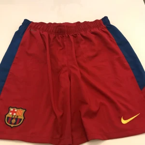 Röda Barcelona shorts - Säljer ett par röda Barcelona shorts som är köpta på camp now. De har en elastisk midja och är perfekta för fotbollsträning. Shortsen har blå detaljer på sidorna och klubbens emblem på ena benet. De är i bra skick och redo för nya äventyr på planen! ⚽️