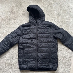 Svart pufferjacka från Jack & Jones - Säljer en svart pufferjacka från Jack & Jones med huva och dragkedja framtill. Jackan har två sidofickor med dragkedja och quiltad design. Perfekt för kyliga dagar och enkel att matcha med olika outfits.