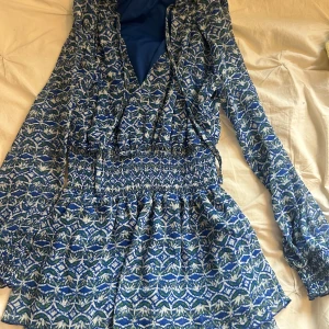 Blå mönstrad kortklänning från Zara - Supersöt blå kortklänning från Zara med långärmade ärmar och elastisk midja. Klänningen har ett unikt mönster i blått, vitt och grönt samt v-ringad omlottfront. Perfekt för dig som gillar färg och snygga detaljer!