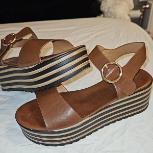Bruna platåsandaler från Catwalk - Snygga bruna sandaler från Catwalk med breda remmar i skinnimitation och stora guldfärgade spännen. Platåsulan har coola svarta och beige ränder som ger extra höjd och stil. Perfekta för dig som vill sticka ut med en trendig look. Ny pris 600kr, använda 2 gånger.