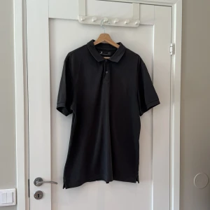 Svart pikétröja från Dressmann 2XL - Klassisk svart pikétröja från Dressmann i storlek 2XL. Tröjan har korta ärmar, krage och två knappar framtill. Perfekt basic-plagg med stilren look, tillverkad i mjuk bomull som känns skön mot huden.