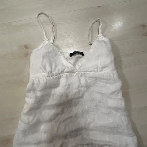 Vit linnetopp från Brandy Melville - Supersöt vit linnetopp från Brandy Melville med smala axelband och v-ringning. Toppen har knyt i ryggen för extra detalj och är gjord i ett luftigt linnematerial, perfekt för varma dagar. Enkel och stilren design som är lätt att matcha.